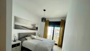 Ein Schlafzimmer mit Bett, Nachttischen, einer Lampe, einem Gemälde und Blick durch ein Fenster.