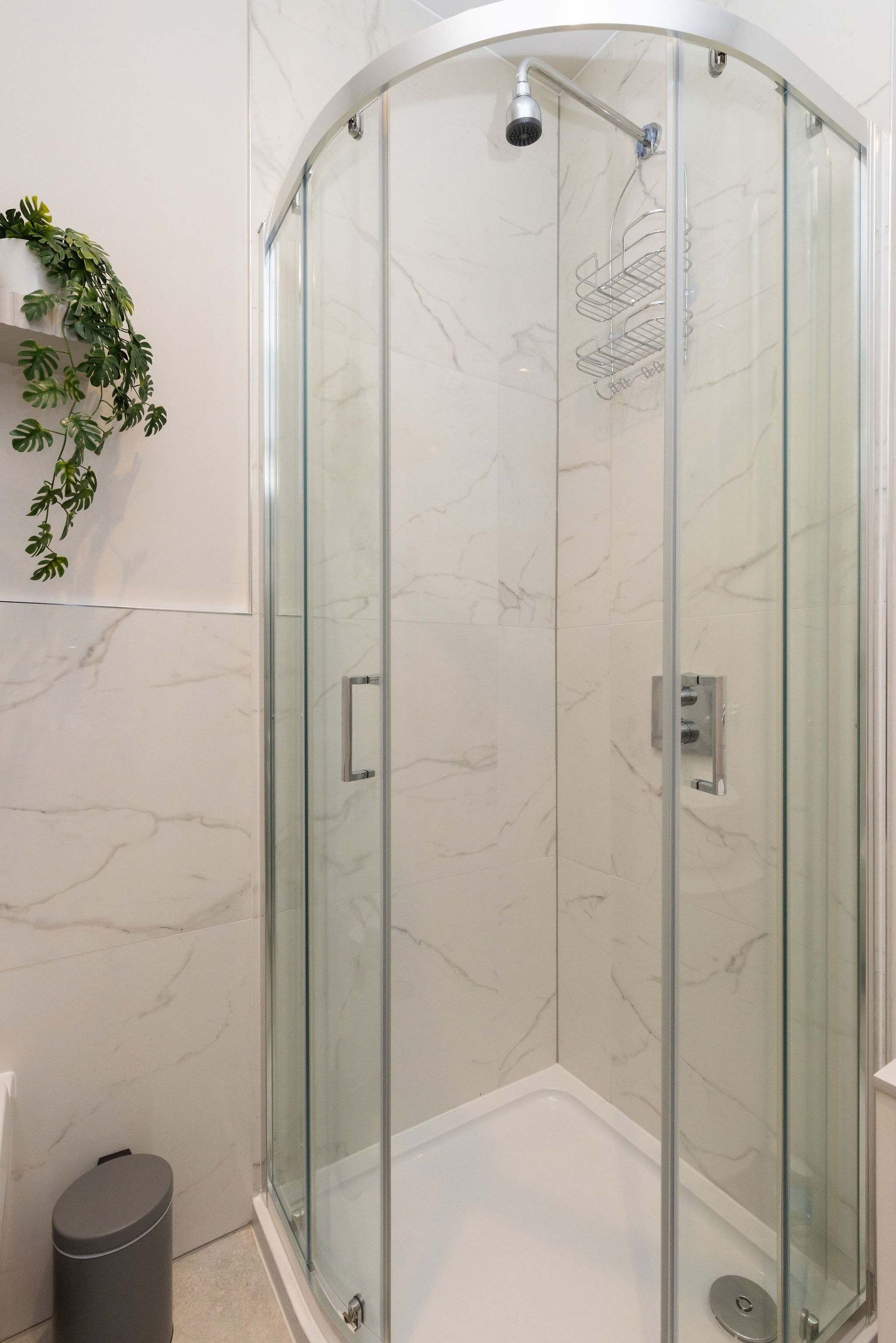 A glass-enclosed shower with a marble wall, a small trash bin, and a potted plant.