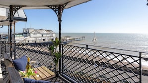 A balcony with a view of the sea, a wooden table with a bottle and glass, and a potted plant.