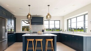 A modern kitchen with dark cabinets, a central island with stools, stainless steel appliances, and large windows offering a view. - Chic Harbour Retreat for 10 With Pool (Chichester)