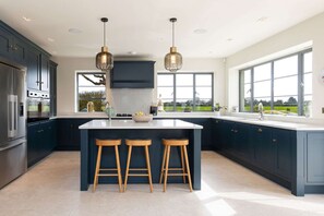 A modern kitchen with dark cabinets, a central island with stools, stainless steel appliances, and large windows offering a view. - Chic Harbour Retreat for 10 With Pool (Chichester)