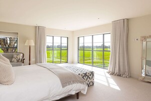 A bedroom with a large bed, a chair, a lamp, a mirror, and a view of a green landscape through large windows.