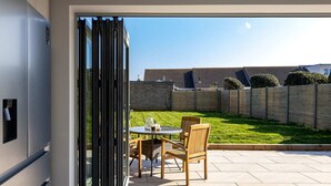 Une terrasse avec une table et des chaises, une porte-fenêtre coulissante et une vue sur une cour arrière avec une clôture et de la pelouse.