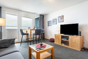 Una sala moderna con un sofá, una mesa de centro, un televisor sobre un mueble de madera, una zona de comedor con sillas y obras de arte en la pared.