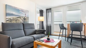 Een moderne hotelkamer met een grijze bank, een klein houten tafeltje, een lamp en een schilderij van een vuurtoren.