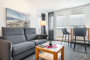 Habitación de hotel moderna con un sofá gris, una pequeña mesa de madera, una lámpara y un cuadro de un faro.