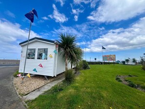 A small, single-story building with a flag on top, surrounded by grass and a palm tree. - Seaside Caravan in a Leisure Park with WiFi & Parking - 3 bedrooms (Rhyl)