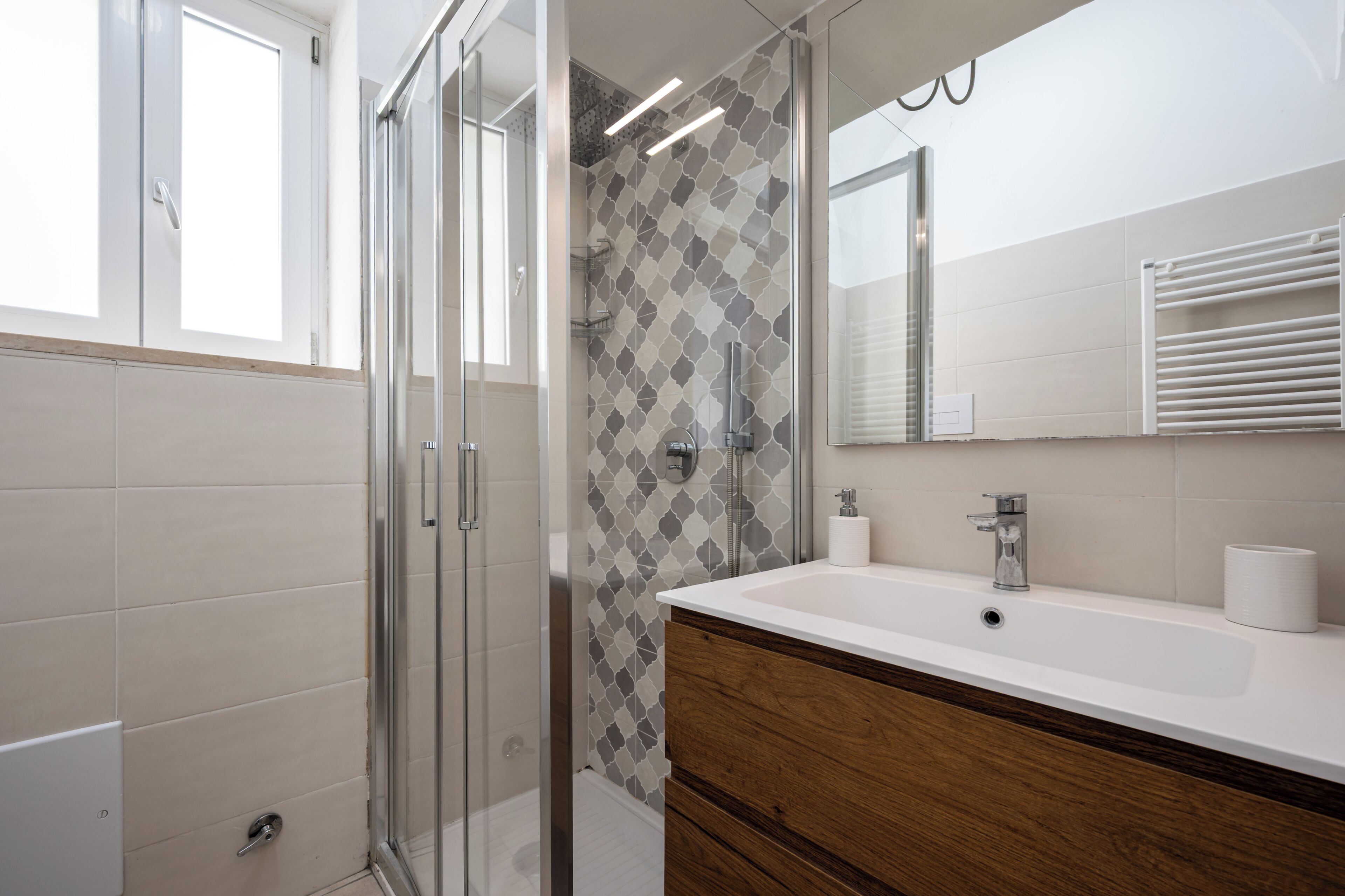 A modern bathroom with a glass shower enclosure, a white sink with a wooden vanity, and a patterned tile backsplash.