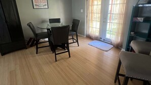 Una habitación con una mesa redonda, cuatro sillas, una estantería, una puerta y una ventana con cortinas.