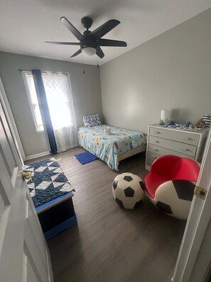 A bedroom with a bed, a desk, a chair, a ceiling fan, a window, and a door.