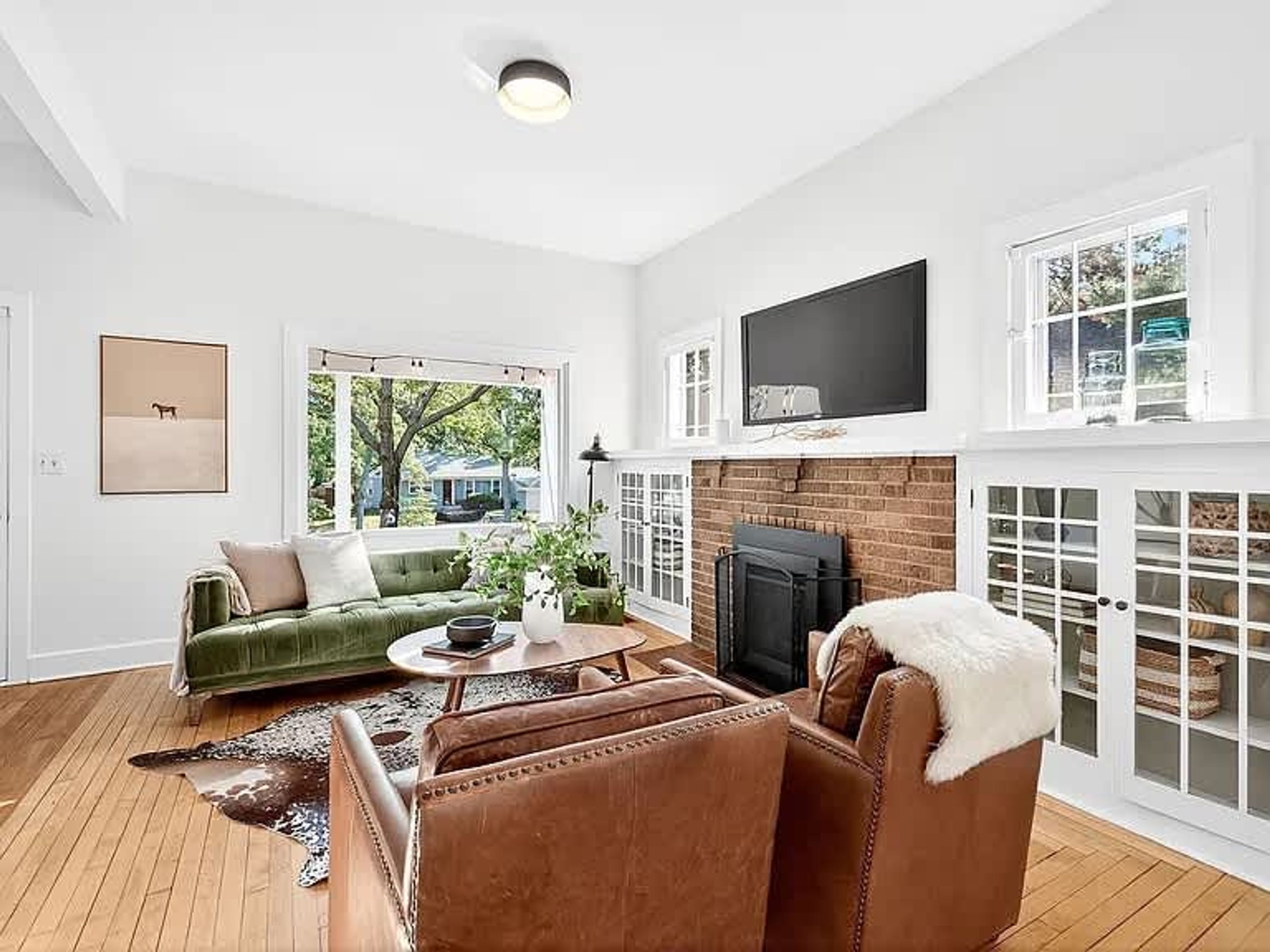 A living room with a green sofa, brown leather chairs, a fireplace, a flat-screen TV, and a large window with a view of trees.