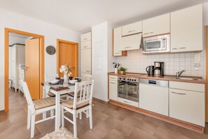 A modern kitchen with white cabinets, a microwave, an oven, and a sink. There is a dining table with chairs and a clock on the wall. - Cozy vacation apartment in Trassenheide, only 150 m from the beach (Trassenheide)