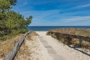 Een zandpad dat naar een strand leidt, met een houten omheining aan één kant en een helderblauwe lucht erboven.