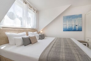 A bedroom with a large bed, a window with curtains, a painting of sailboats, and a bedside table. - Cozy vacation apartment in Trassenheide, only 150 m from the beach (Trassenheide)