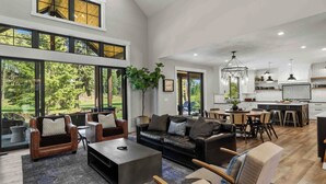 A spacious living area with a black leather sofa, multiple armchairs, and a dining table. Large windows offer views of a lush garden. - Memories Made on Prospector's 16th Fairway-Spa & Pets (Cle Elum)