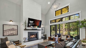 A modern living room with a fireplace, comfortable seating, a large TV, and large windows offering a view of the outdoors. - Memories Made on Prospector's 16th Fairway-Spa & Pets (Cle Elum)
