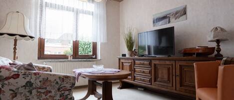 A living room with a sofa, a table, a TV on a wooden cabinet, and a window with curtains.