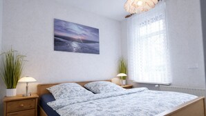 A bedroom with a bed, two bedside tables with lamps, a window with sheer curtains, and a wall-mounted painting. - Your perfect time-out in Duhnen - cozy, central and close to the beach (Cuxhaven)