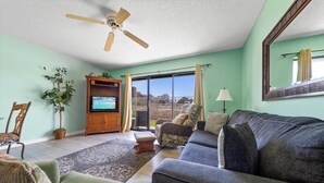 Ein Wohnzimmer mit einer Couch, Sesseln, einem Fernseher, einem Deckenventilator, einer Pflanze und Blick durch eine Glastür.