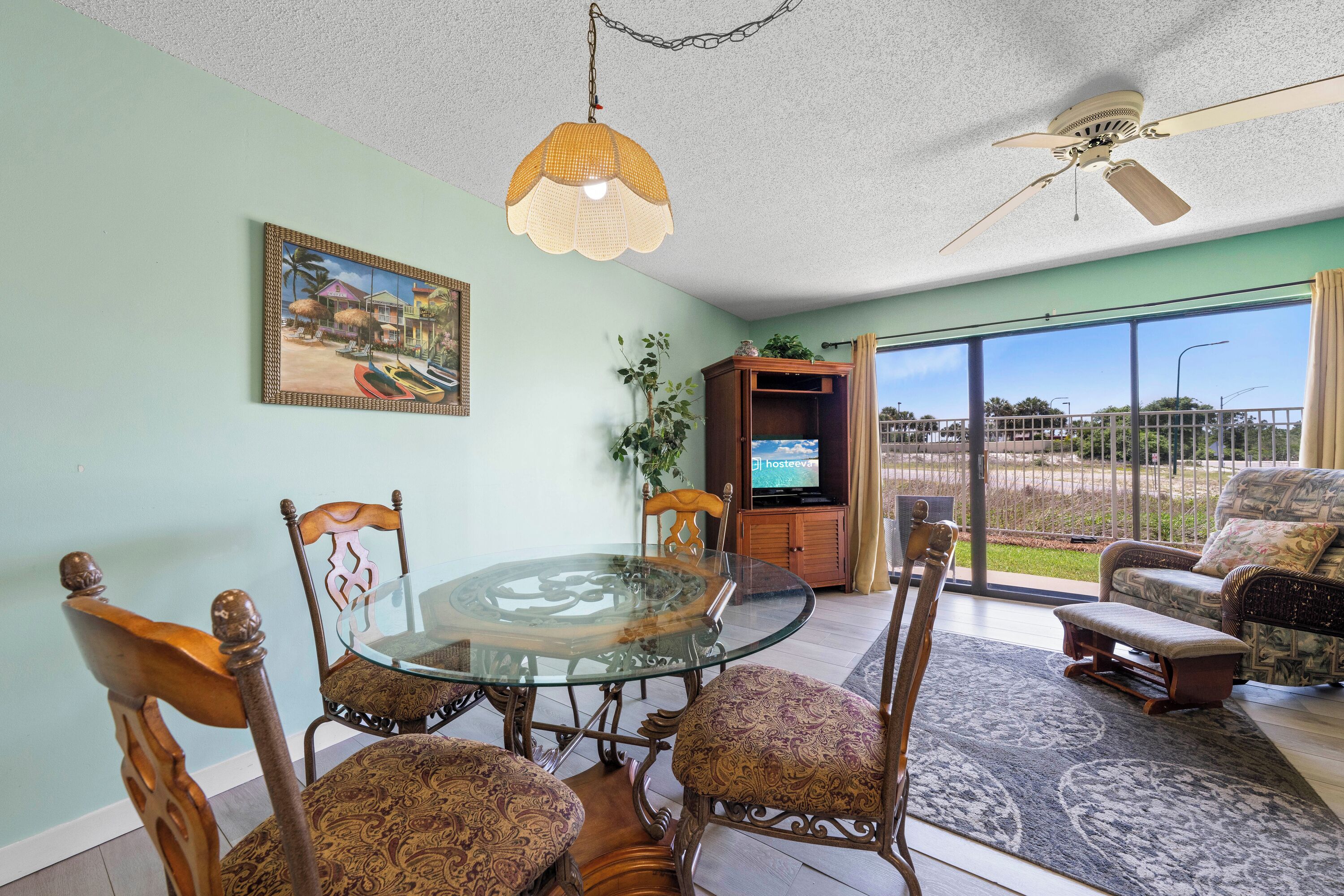 A dining area with a glass table, chairs, a ceiling fan, a television, and a view through a glass door.
