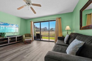 A living room with a ceiling fan, a television, a sofa, a chair, a lamp, and a view of the outdoors through a glass door.