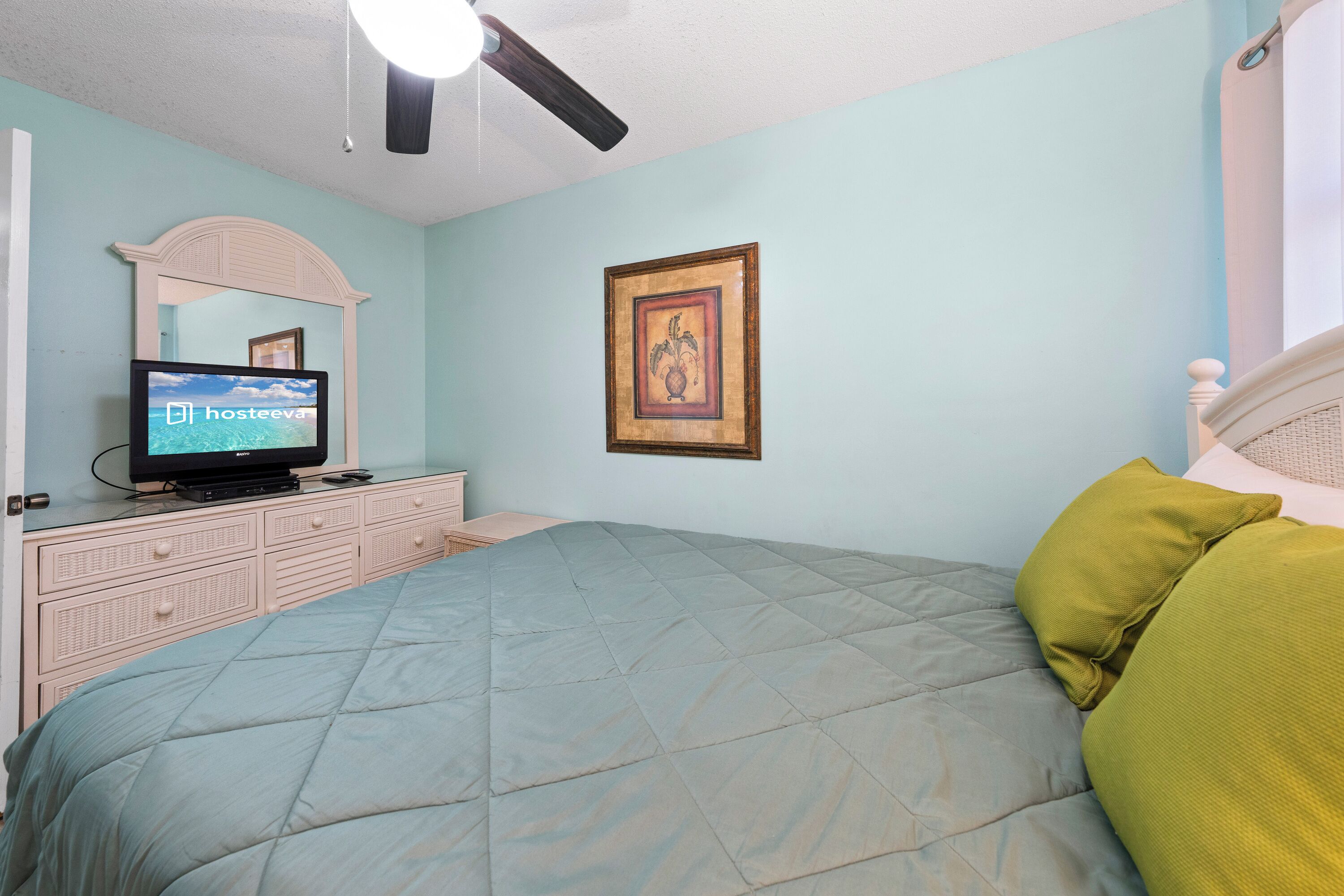 A bedroom with a bed, a dresser, a television, a ceiling fan, and a framed picture on the wall.