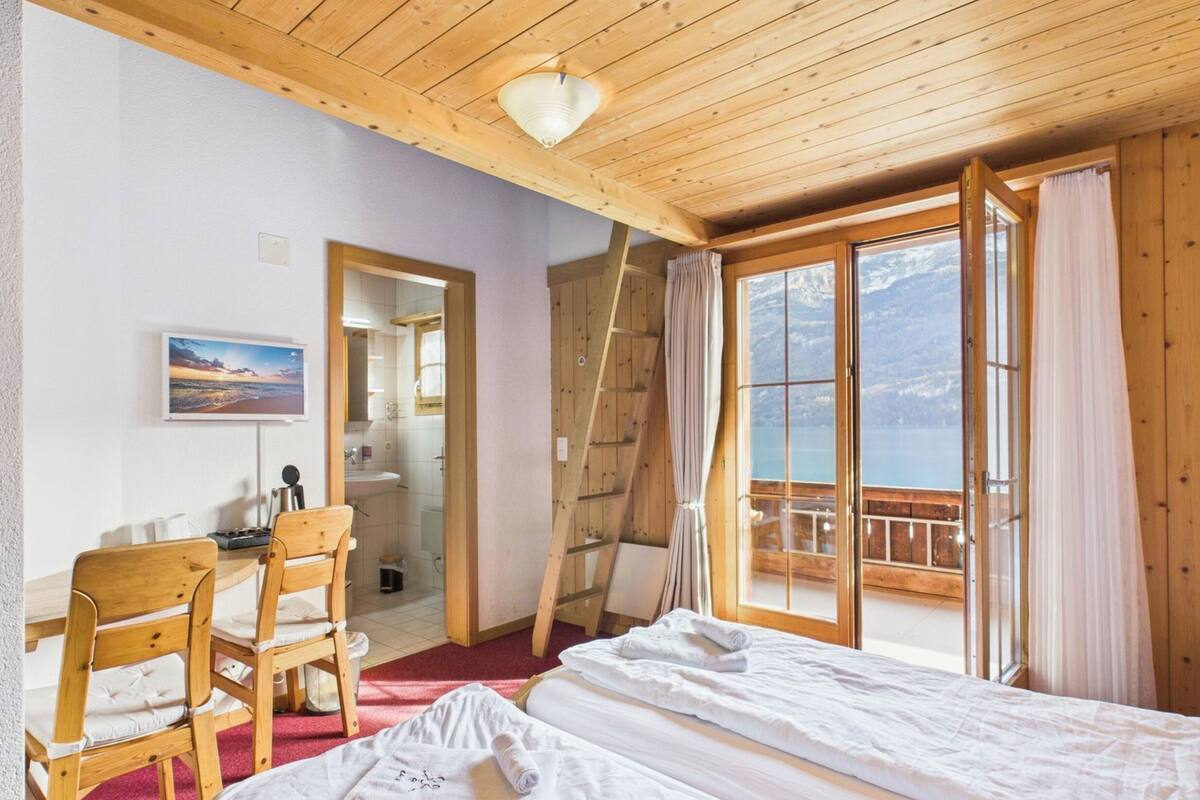 A wooden cabin room with a bed, chairs, a desk, a bathroom, and a balcony with a view.