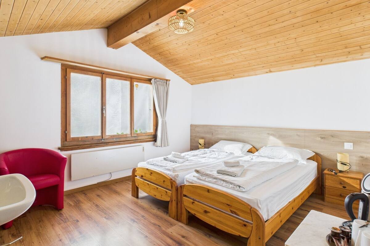 A bedroom with a wooden ceiling, a bed with white linens, a red chair, and a window with curtains.