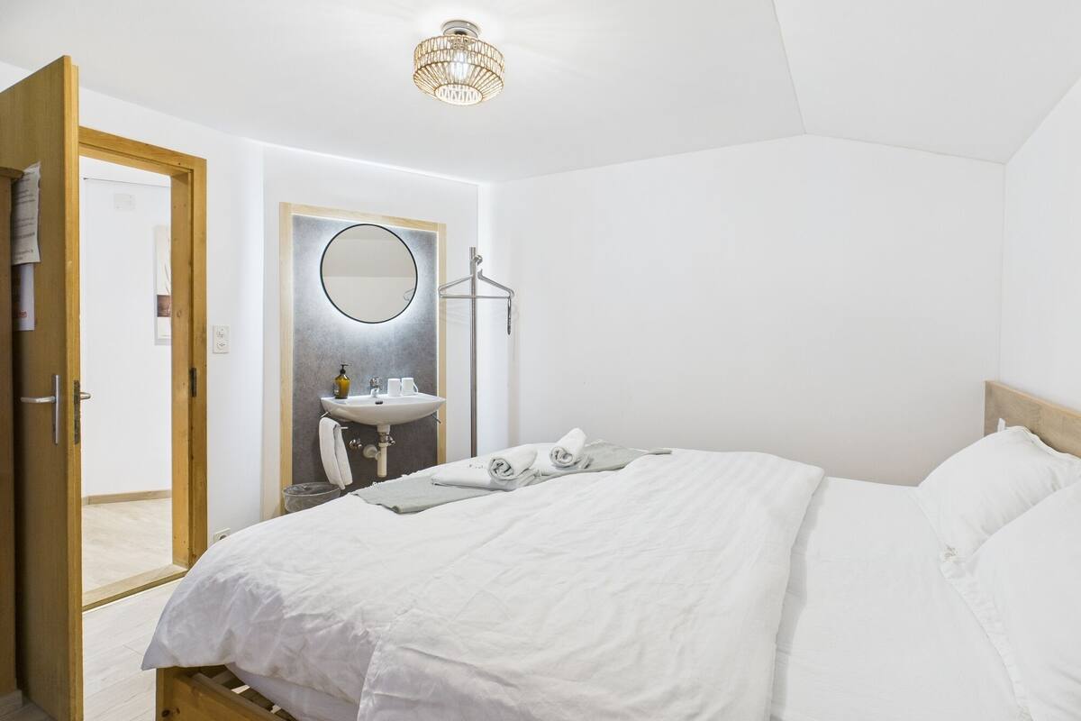 A bedroom with a bed, a bedside table, a mirror, a hanging light fixture, and a door leading to another room.