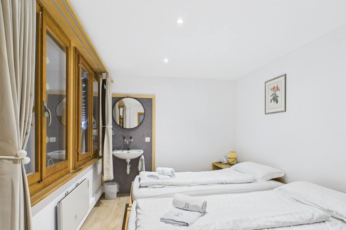 A room with two beds, a wooden-framed window, a round mirror, a sink, and a framed picture on the wall.