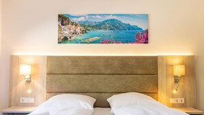 A hotel room with a wooden headboard, two bedside tables with lamps, a bed with white linens, and a painting of a coastal landscape on the wall.