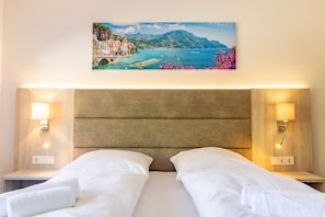 A hotel room with a wooden headboard, two bedside tables with lamps, a bed with white linens, and a painting of a coastal landscape on the wall.