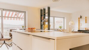 A modern kitchen with a large island, a dining area with chairs, a bookshelf, and a view through the window.