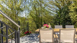 An outdoor patio with a wooden table and chairs, surrounded by trees and a metal fence. - Panoramic view of Lake Edersee - View. Balcony. Sauna. Top location. (Hermfurth-Edersee)