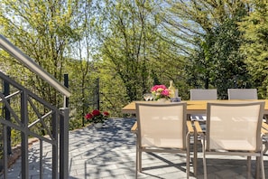 An outdoor patio with a wooden table and chairs, surrounded by trees and a metal fence.