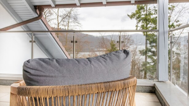 Een rieten stoel met een grijze kussen, een glazen balustrade en uitzicht op bomen en bergen.