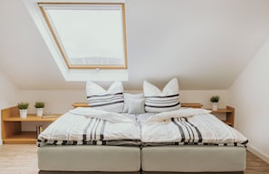 Une chambre avec une lucarne, un lit avec une literie à rayures, deux tables de chevet ornées de plantes et une étagère en bois.