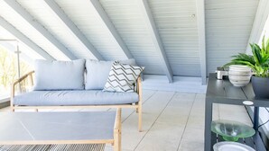 Een overdekt terras met een bank, een tafel, een potplant en een stapel borden.