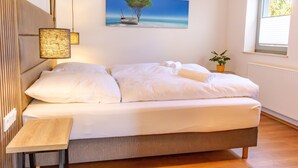 Uma cama bem arrumada com lençóis brancos, uma mesa de cabeceira, um quadro na parede de uma árvore e um barco, e uma janela com persianas.