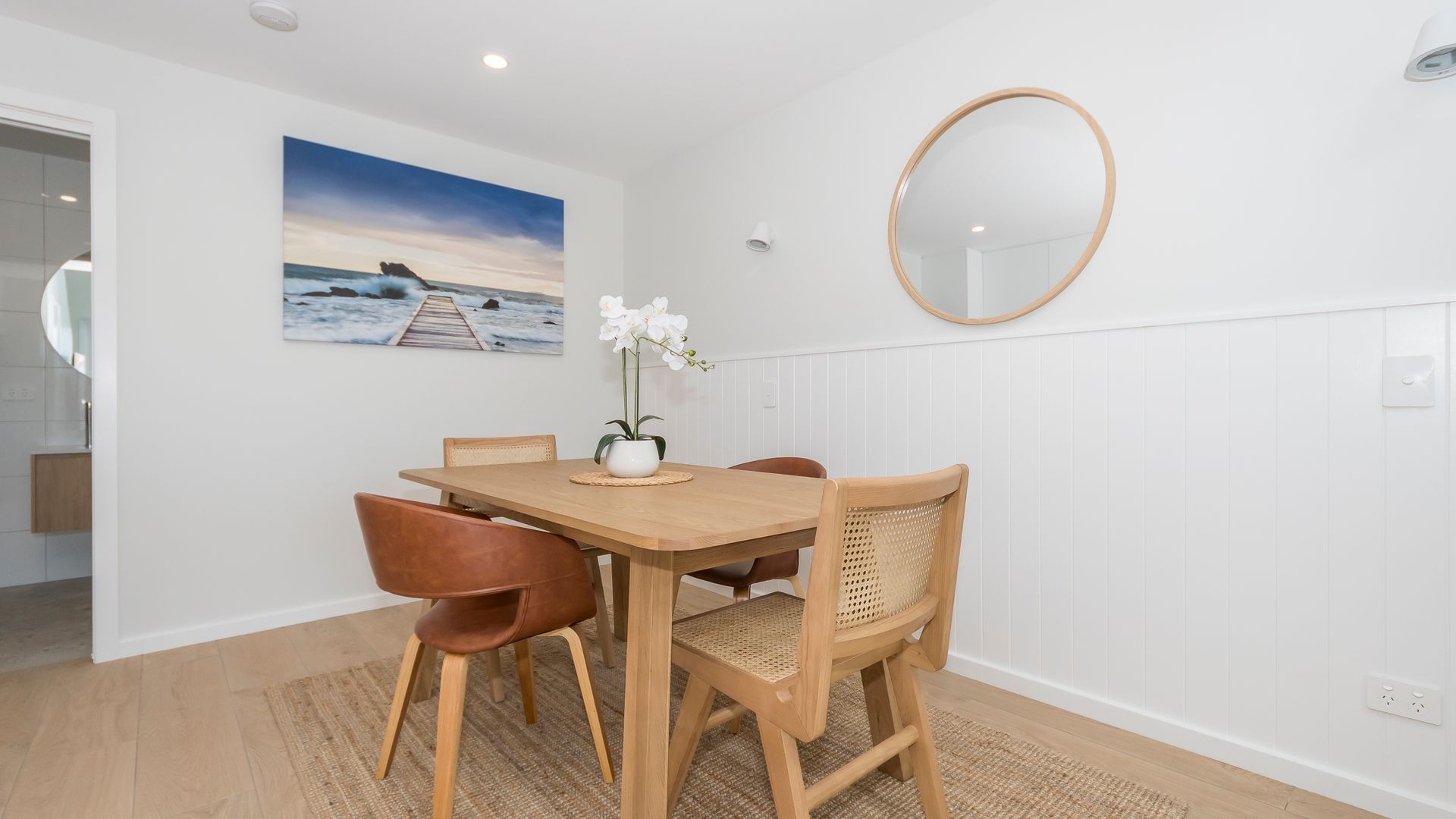 A dining area with a wooden table, four chairs, a vase with flowers, a wall-mounted mirror, and a painting of a pier.