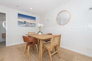 Ein Essbereich mit einem Holztisch und -stühlen, einem Bild einer Strandlandschaft, einem runden Spiegel und einer weißen Wand.