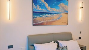 Une chambre à coucher avec un lit, des tables de chevet, un tableau représentant une plage et des luminaires fixés au mur.