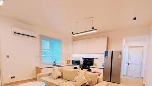 Una sala de estar moderna con un sofá, dos mesitas auxiliares, una zona de cocina con electrodomésticos y una puerta que da a otra habitación.