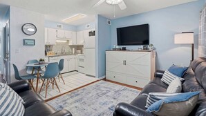 A living room with a black leather couch, a flat-screen TV, a ceiling fan, and a kitchen area with white cabinets and a dining table. - "Dune Delight" At Sea Mystic (North Myrtle Beach)