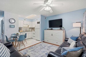 Ein Wohnzimmer mit schwarzem Ledersofa, Flachbildfernseher, Deckenventilator und Küchenzeile mit weißen Schränken sowie einem Esstisch.