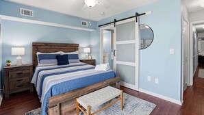 A bedroom with a bed, bedside tables, a bench, a ceiling fan, a mirror, and a door. - "Dune Delight" At Sea Mystic (North Myrtle Beach)