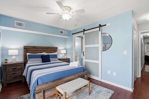 Ein Schlafzimmer mit einem Bett, Nachttischen, einer Bank, einem Deckenventilator, einem Spiegel und einer Tür.