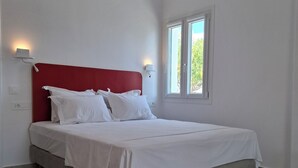 Un dormitorio con cama, ropa de cama blanca, cabecera roja, dos lámparas fijadas a la pared, una ventana y un enchufe.