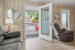 Een woonkamer met een grijze bank, een fauteuil, een ronde spiegel en een lamp. Een houten wandpaneel en een deur naar buiten zijn zichtbaar.