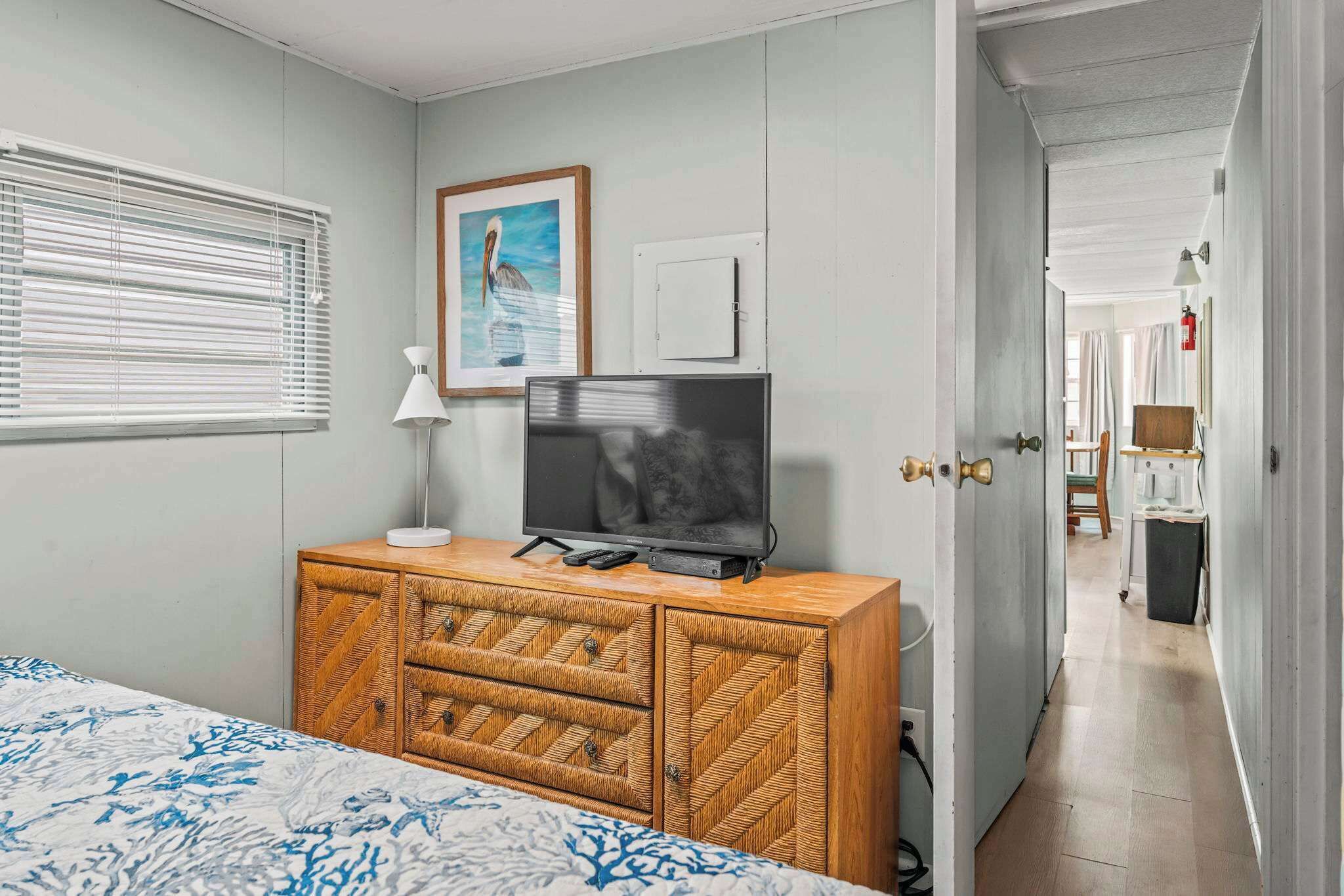 A bedroom with a bed, a television on a wooden dresser, a painting on the wall, and a hallway leading to another room.